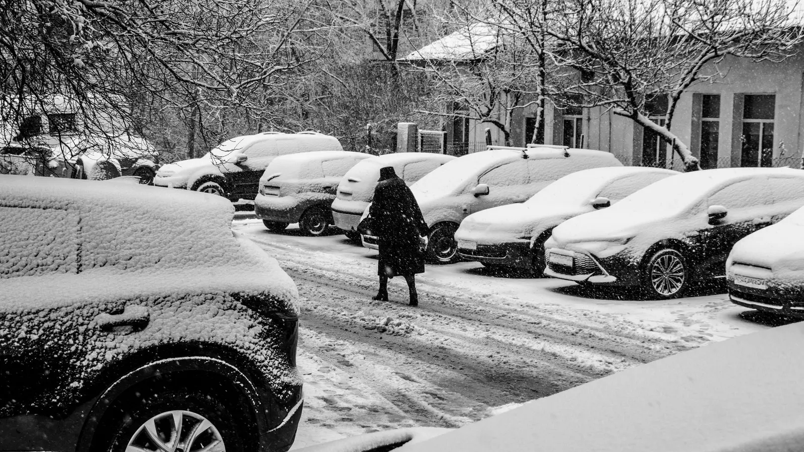 Person walking through a snowy parking lot past snow-covered cars