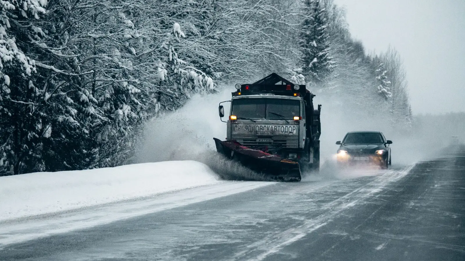 Plow truck clearing a wooded country road with a car following behind